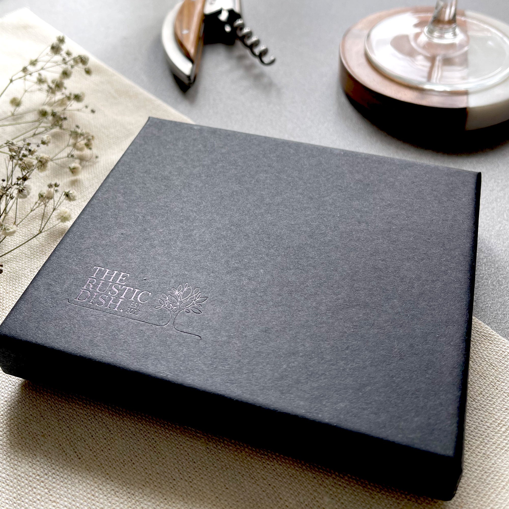 Dark gray box with 'The Rustic Dish' logo on a light surface with wine-related items.