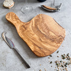 A rustic olive wood cheese board, displayed alongside a cheese knife, bottle of wine, cork screw, and a small round dish of what appears to be crackers. 