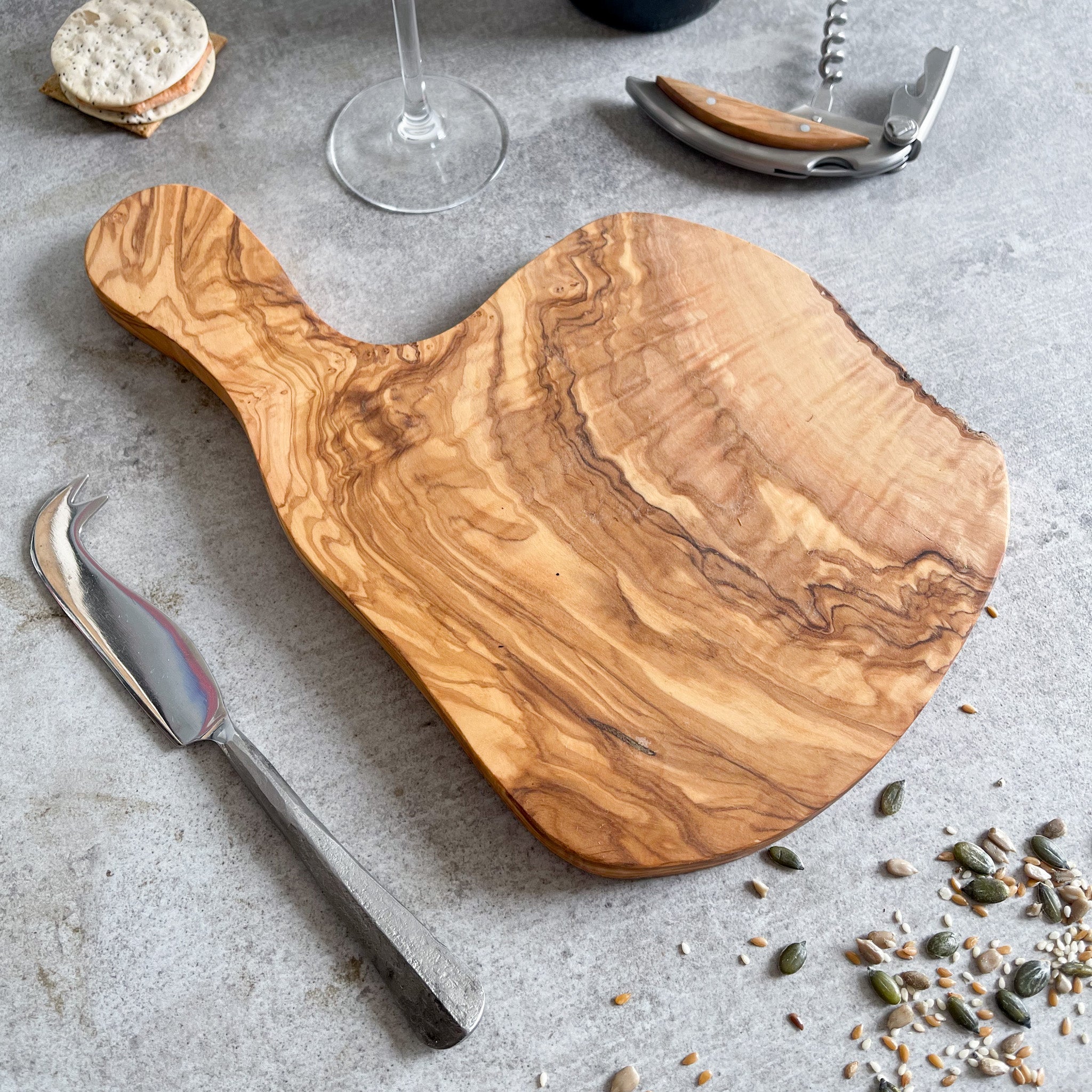 A rustic olive wood cheese board, displayed alongside a cheese knife, bottle of wine, cork screw, and a small round dish of what appears to be crackers. 