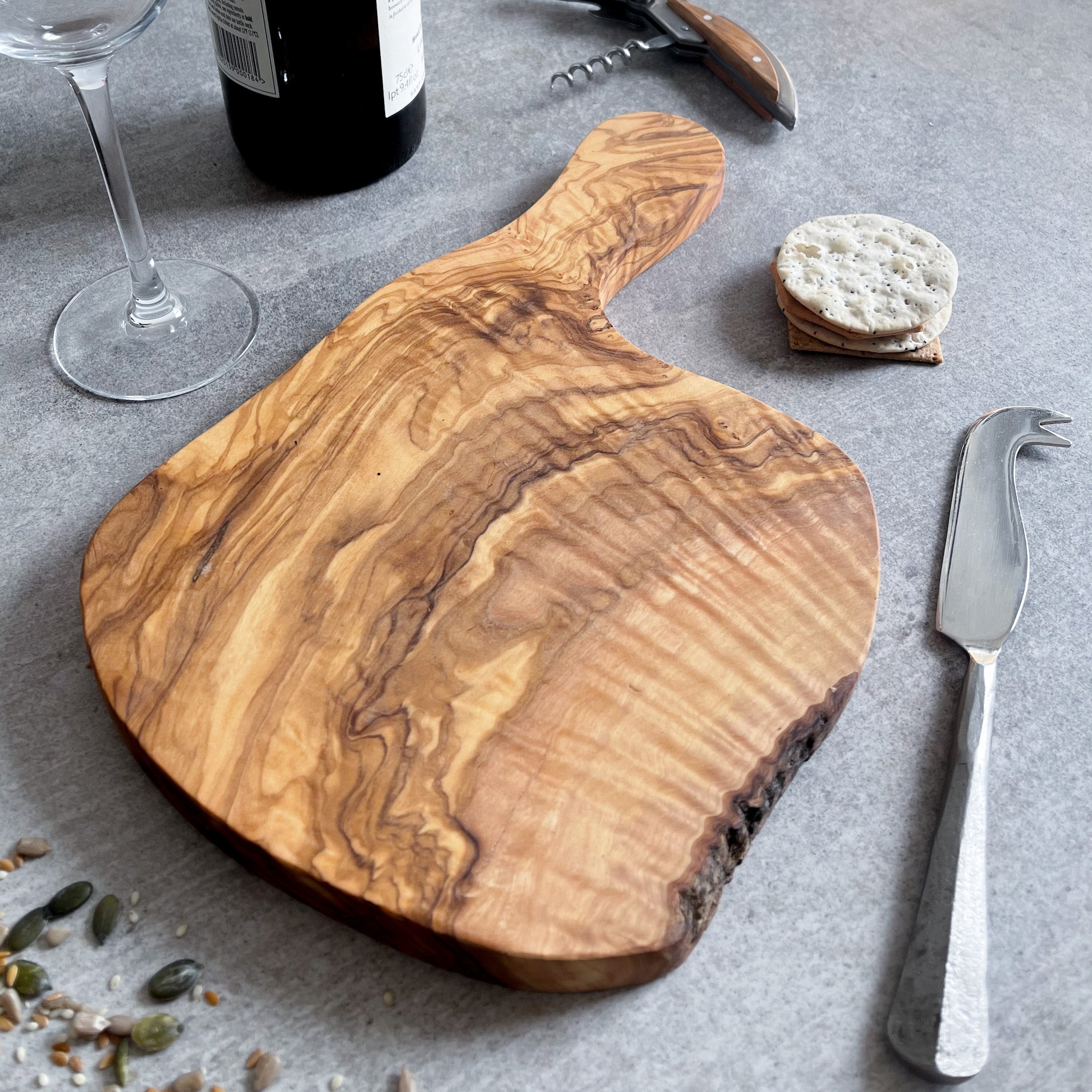 A rustic olive wood cheese board with a cheese knife alongside, displayed alongside a bottle of wine, cork screw, and some crackers. 