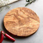a round olive wood cutting board on a grey surface with some chillies and flowers.