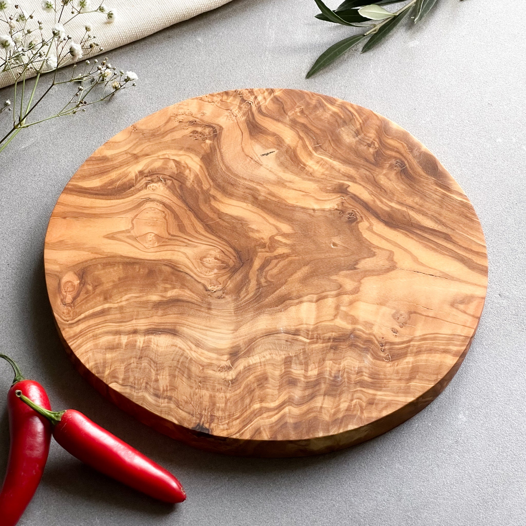a round olive wood cutting board on a grey surface with some chillies and flowers.