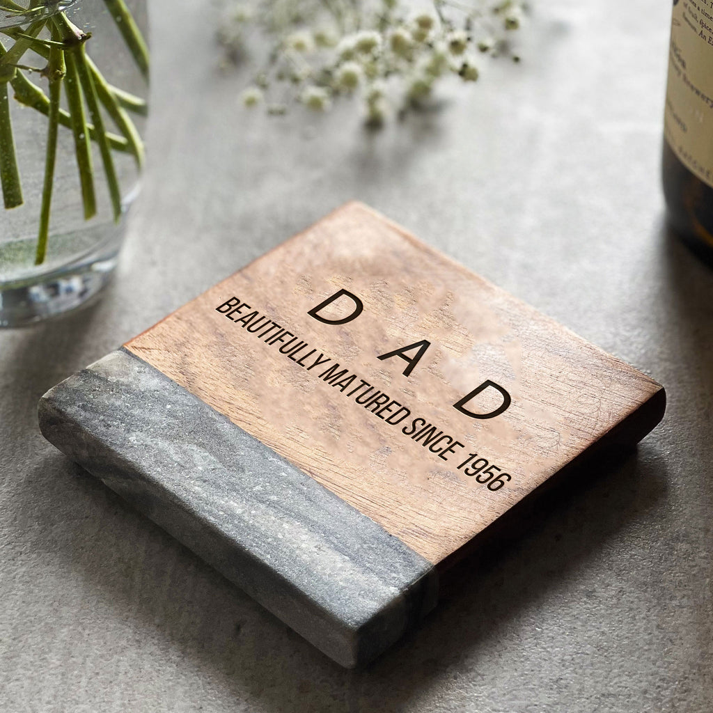 A personalized wooden coaster with the text 'DAD' engraved on it, placed on a table with a glass and some fllowers in the background.