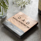 A personalized wooden coaster with the text 'DAD' engraved on it, placed on a table with a glass and some fllowers in the background.