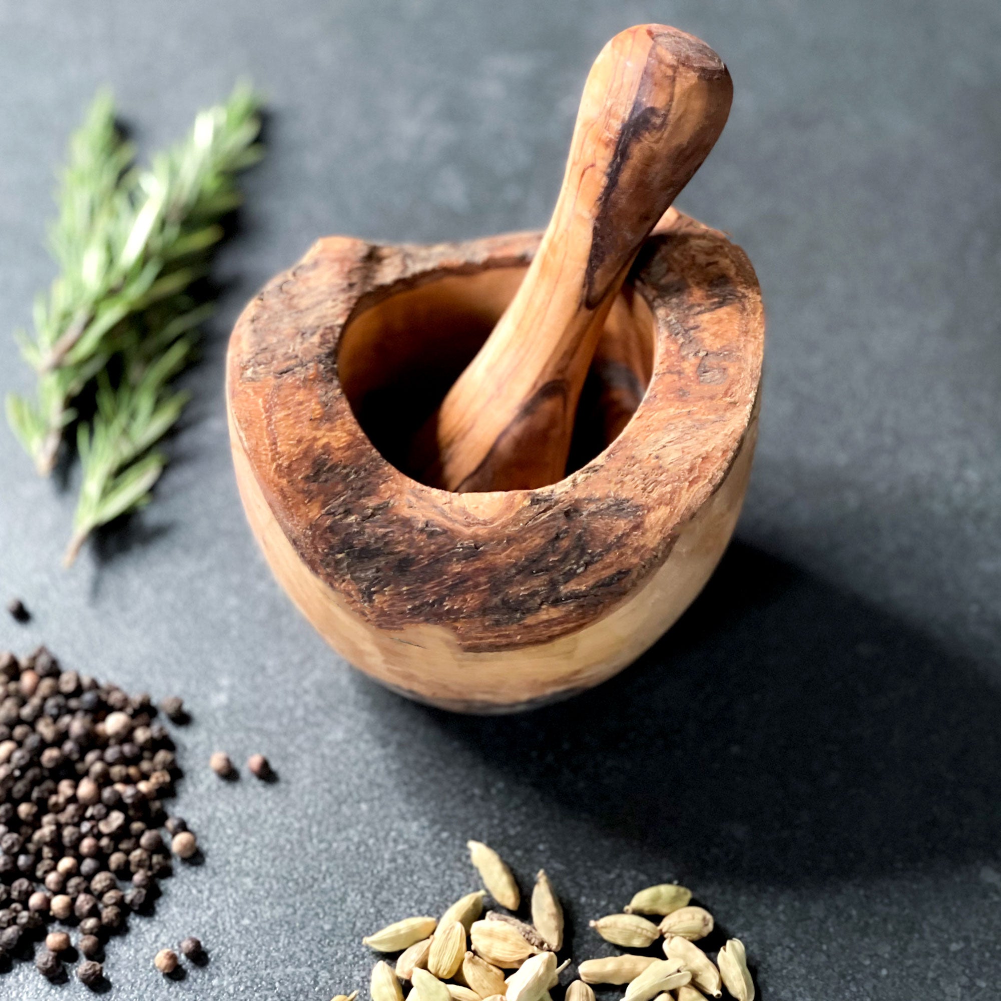 an olive wood pestle and mortar with rustic edge on a dark surface surrounded by herbs and spices.