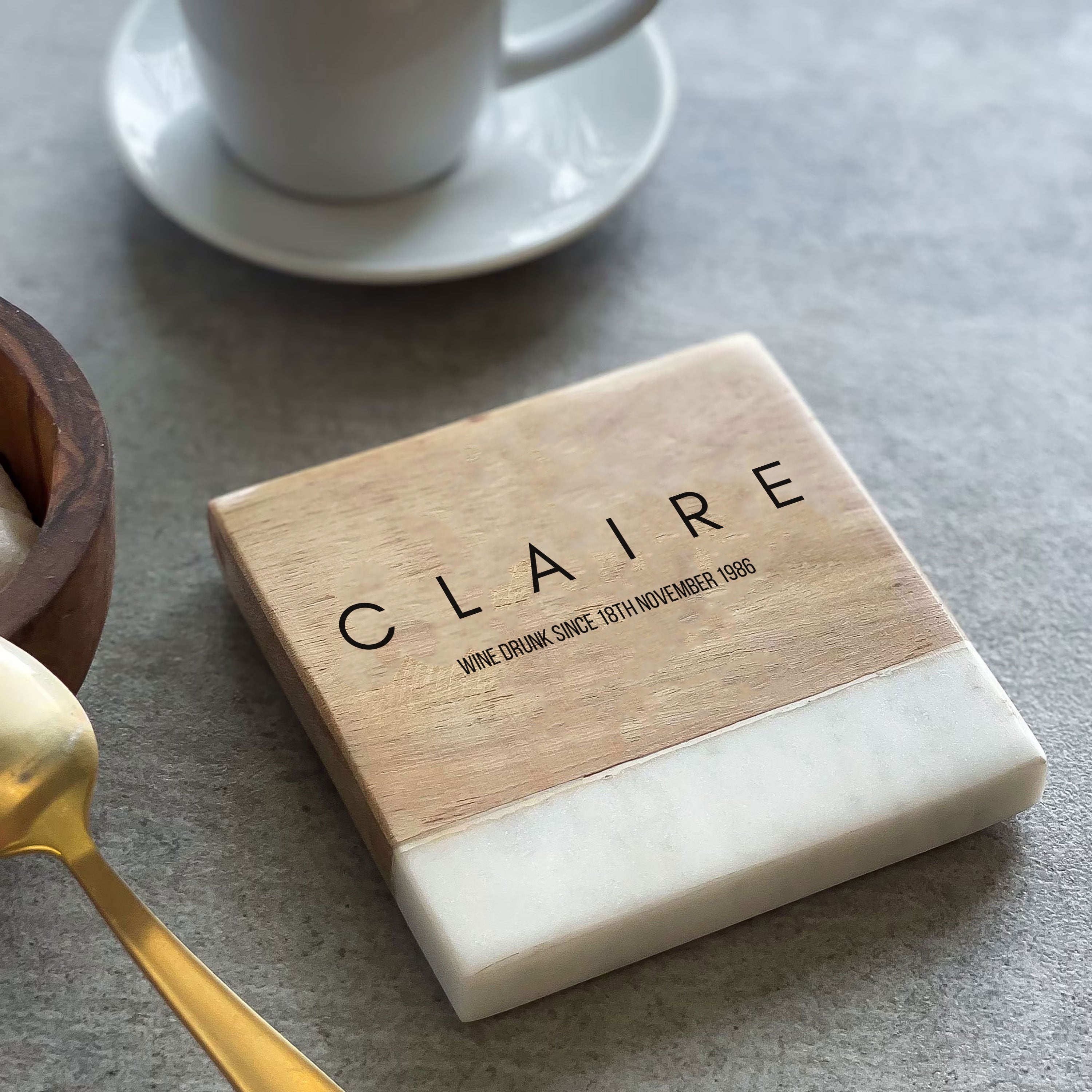 A personalized wooden coaster with the text 'CLAIRE' engraved on it, placed on a table with a cup and saucer in the background along with a sugar pot and spoon