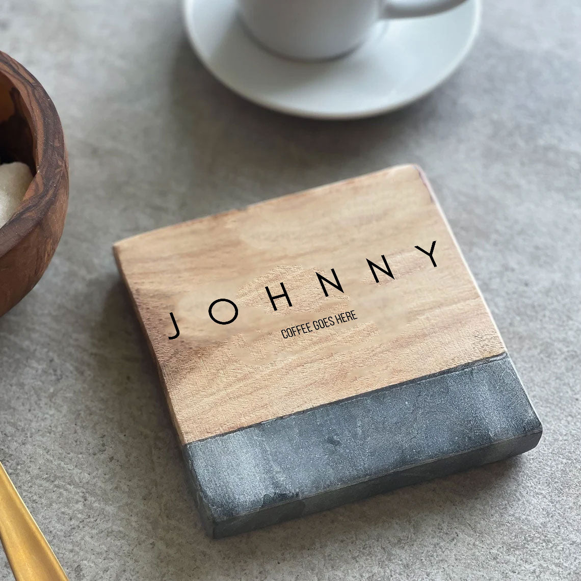 A personalized wooden coaster with the text 'JOHNNY' engraved on it, placed on a table with a cup and saucer in the background along with a sugar pot and spoon