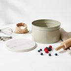 Baking setup with a round cake tin, flour, berries, and a rolling pin on a light surface.