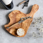A rustic olive wood cheese board with a cheese knife on top, displayed alongside a bottle of wine, cork screw, and a small round dish of what appears to be crackers. Decorative white flowers are also visible to the side.