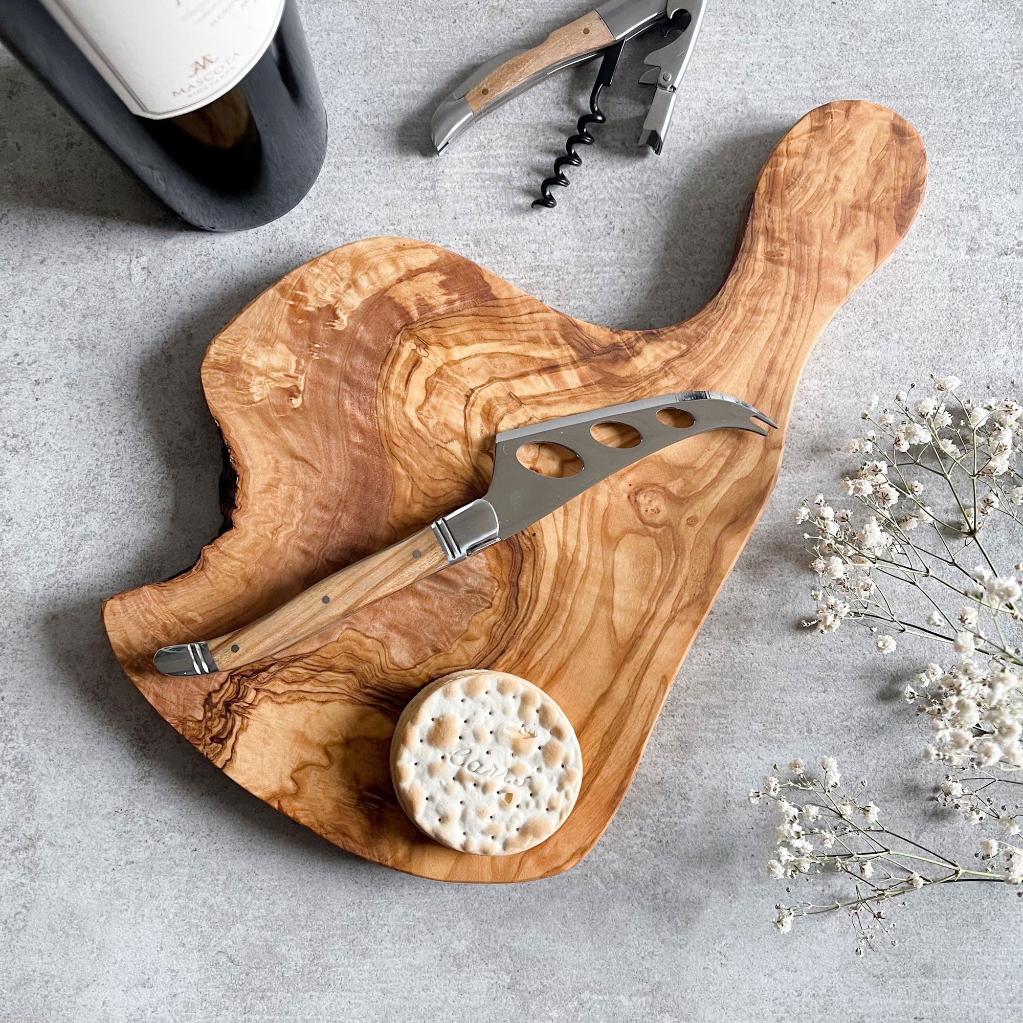 A rustic olive wood cheese board with a cheese knife on top, displayed alongside a bottle of wine, cork screw, and a small round dish of what appears to be crackers. Decorative white flowers are also visible to the side.