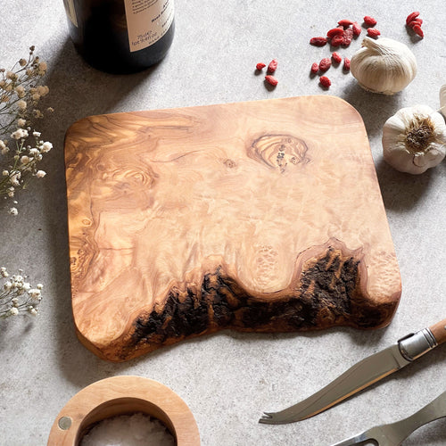 A rustic olive wood cheese board with a handfinished edge, displayed with a bottle of wine, garlic, and a knife.