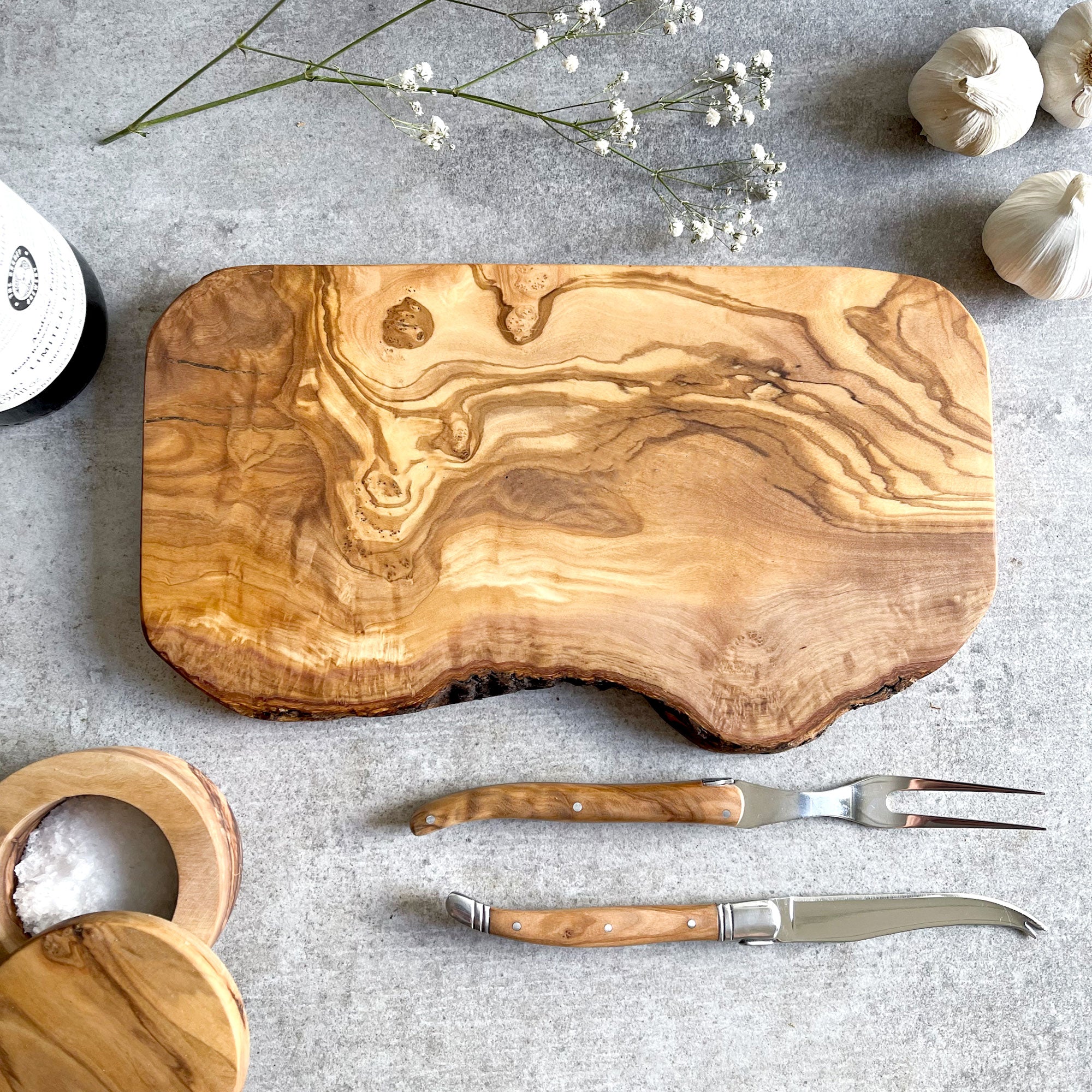 A rustic olive wood cheese/chopping board with a handcrafted appearance, accompanied by wine, garlic and salt. A small knife and serving knife with 
wooden handles are also visible next to the board.