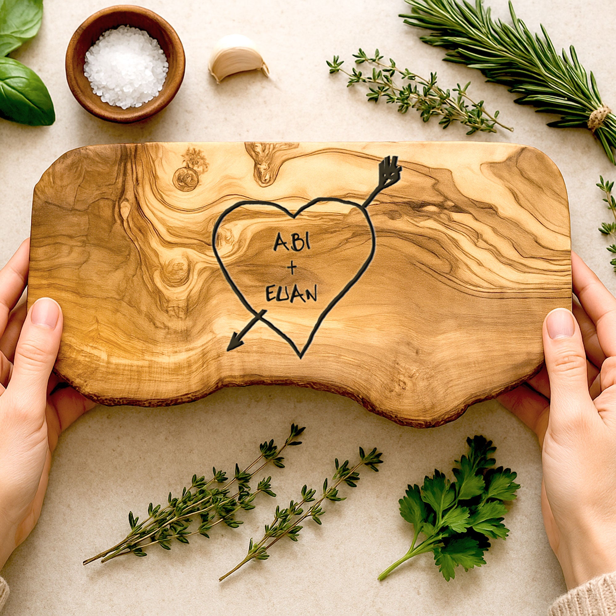 A personalized rustic carved heart cheese board with engraved names, held in hands with a background of herbs and a small bowl.