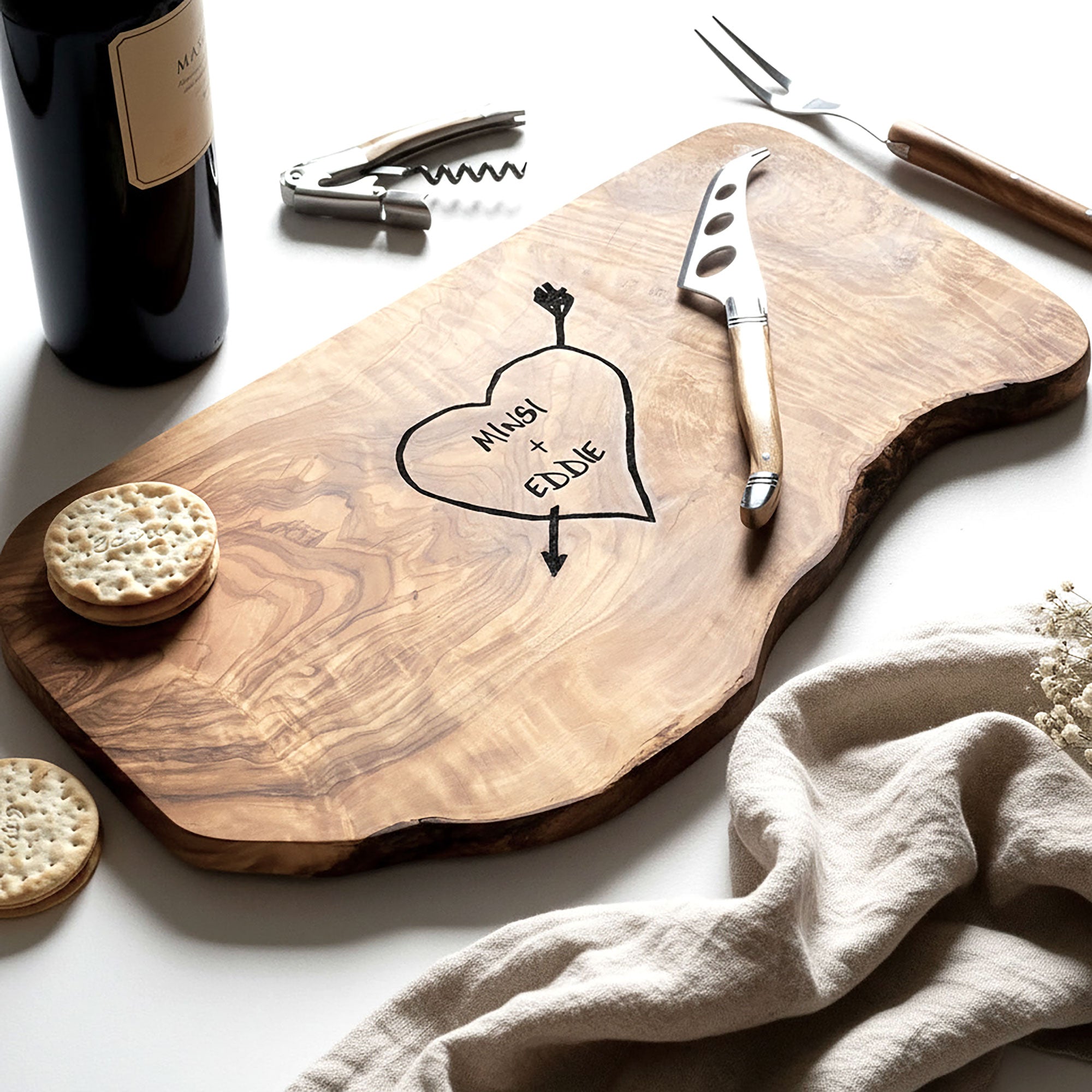 Wooden cutting board with engraved names, knife, crackers, and bottle on a white surface