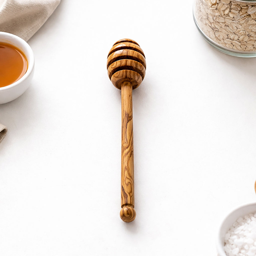 Wooden honey dipper on a white surface with a bowl of tea and jar of oats in the background.