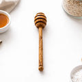 Wooden honey dipper on a white surface with a bowl of tea and jar of oats in the background.