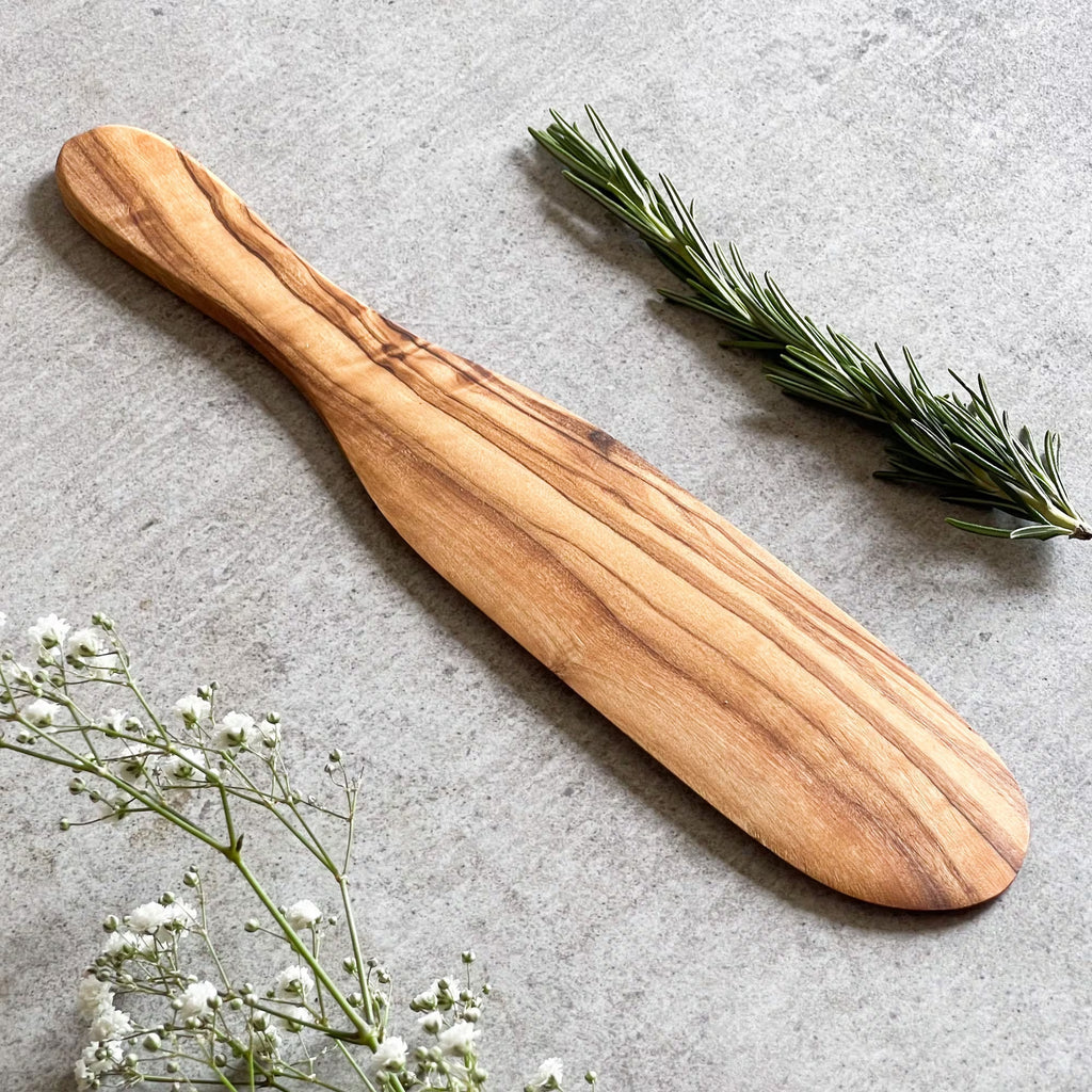 A wooden spatula with a smooth finish and a handle, displayed on a grey surface, accompanied by some green herbs and white flowers.