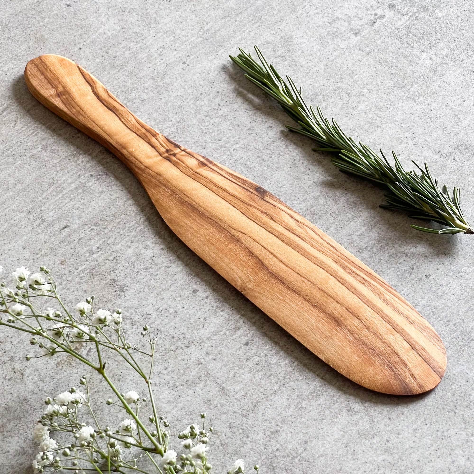 A wooden spatula with a smooth finish and a handle, displayed on a grey surface, accompanied by some green herbs and white flowers.