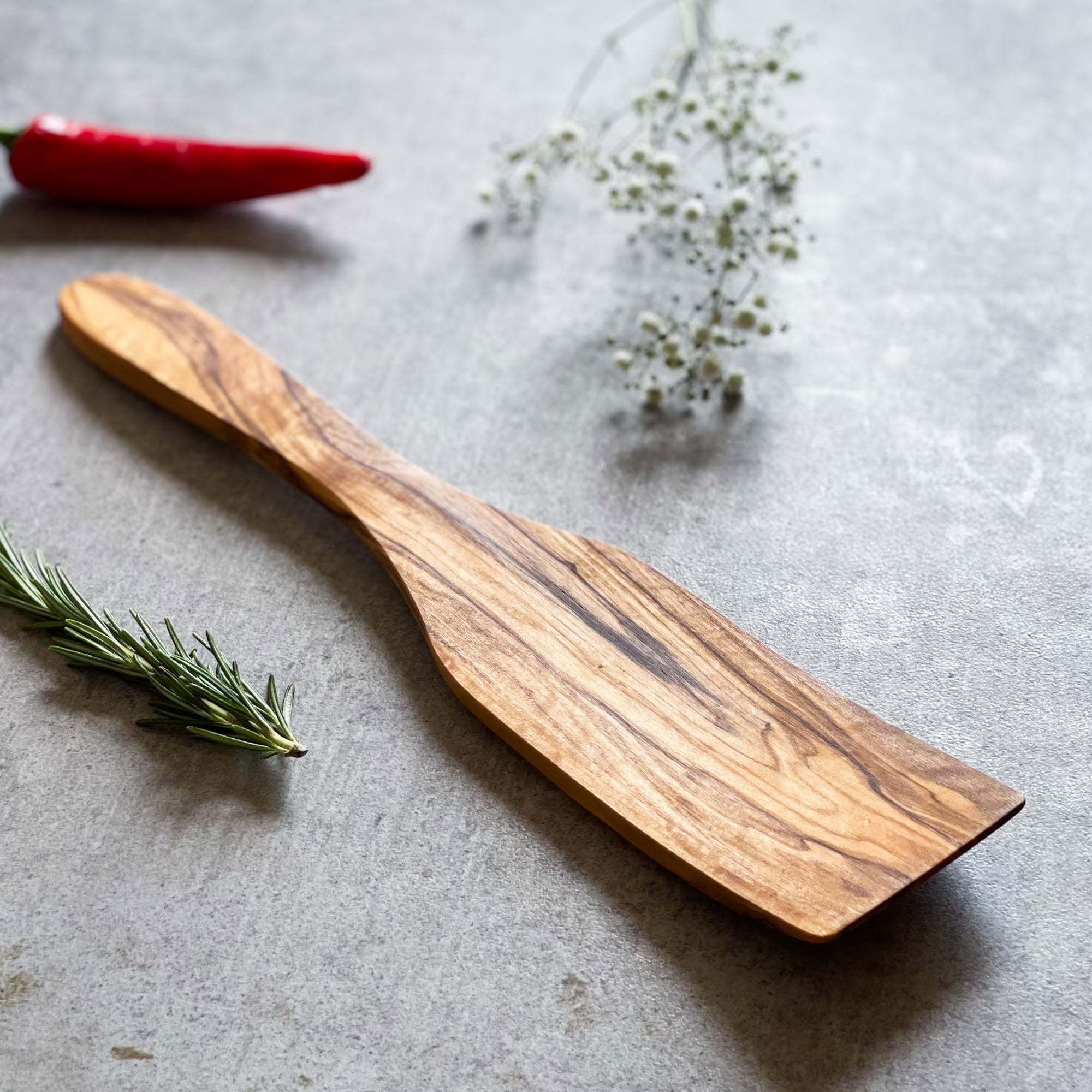 A handcrafted olive wood spatula with a rustic handle, displayed on a grey surface, with a red chili and some herbs in the background.