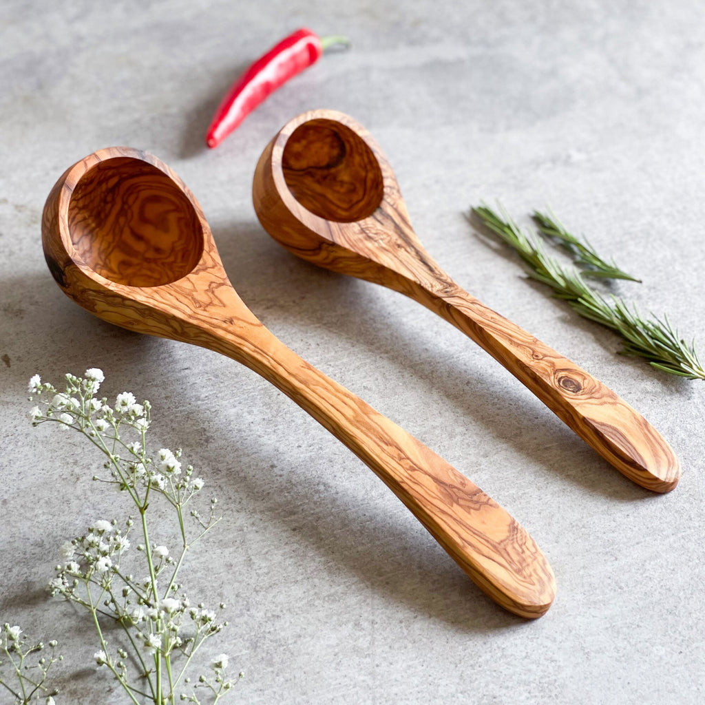Two handcrafted olive wood ladles with distinctive grain patterns, displayed on a grey surface with a red chili and green herbs in the background.