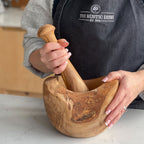 A person holding a brown wooden pestle and mortar, with the pestle resting inside the mortar. The person is wearing a black apron with 'THE RUSTIC DISH' written on it.