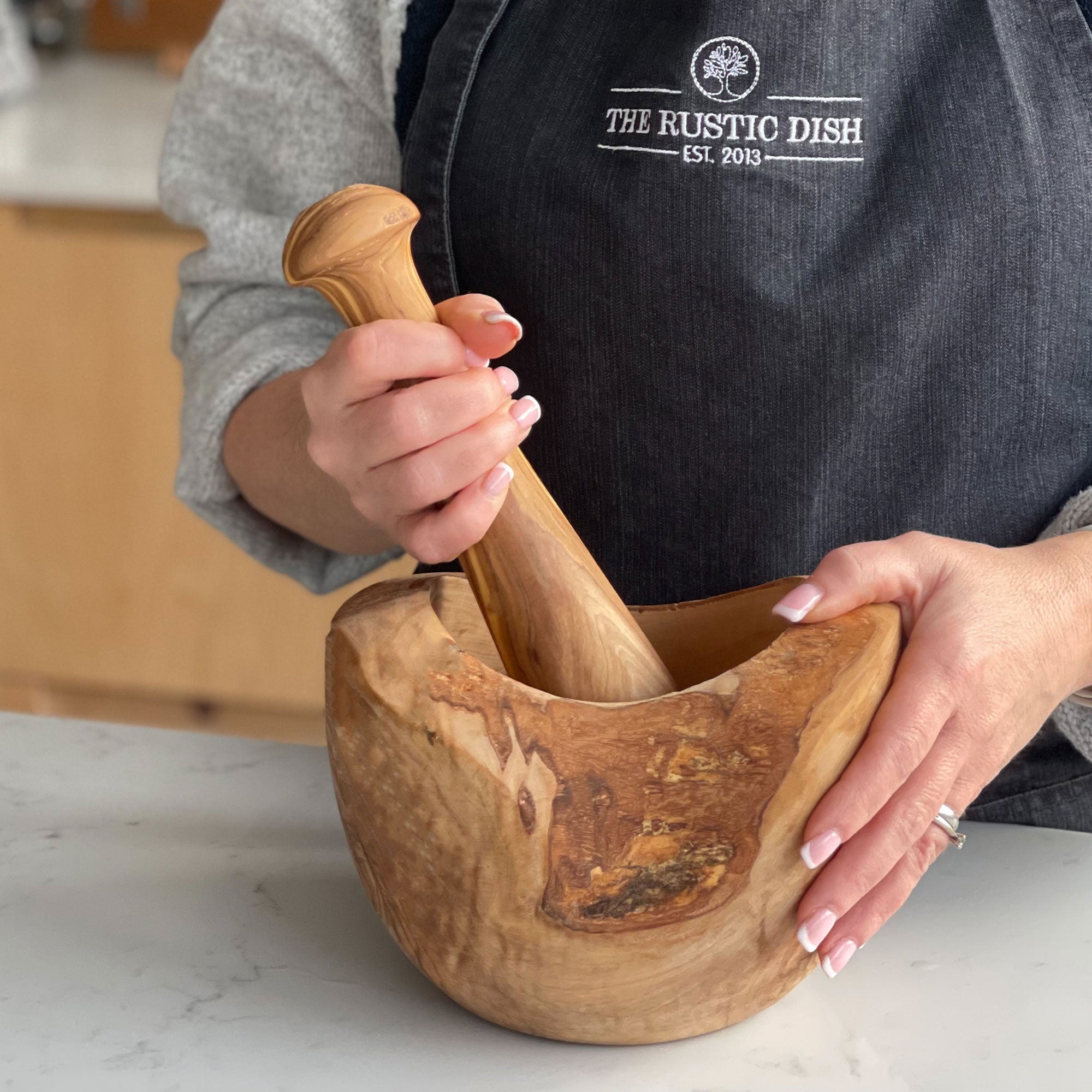 A person holding a brown wooden pestle and mortar, with the pestle resting inside the mortar. The person is wearing a black apron with 'THE RUSTIC DISH' written on it.