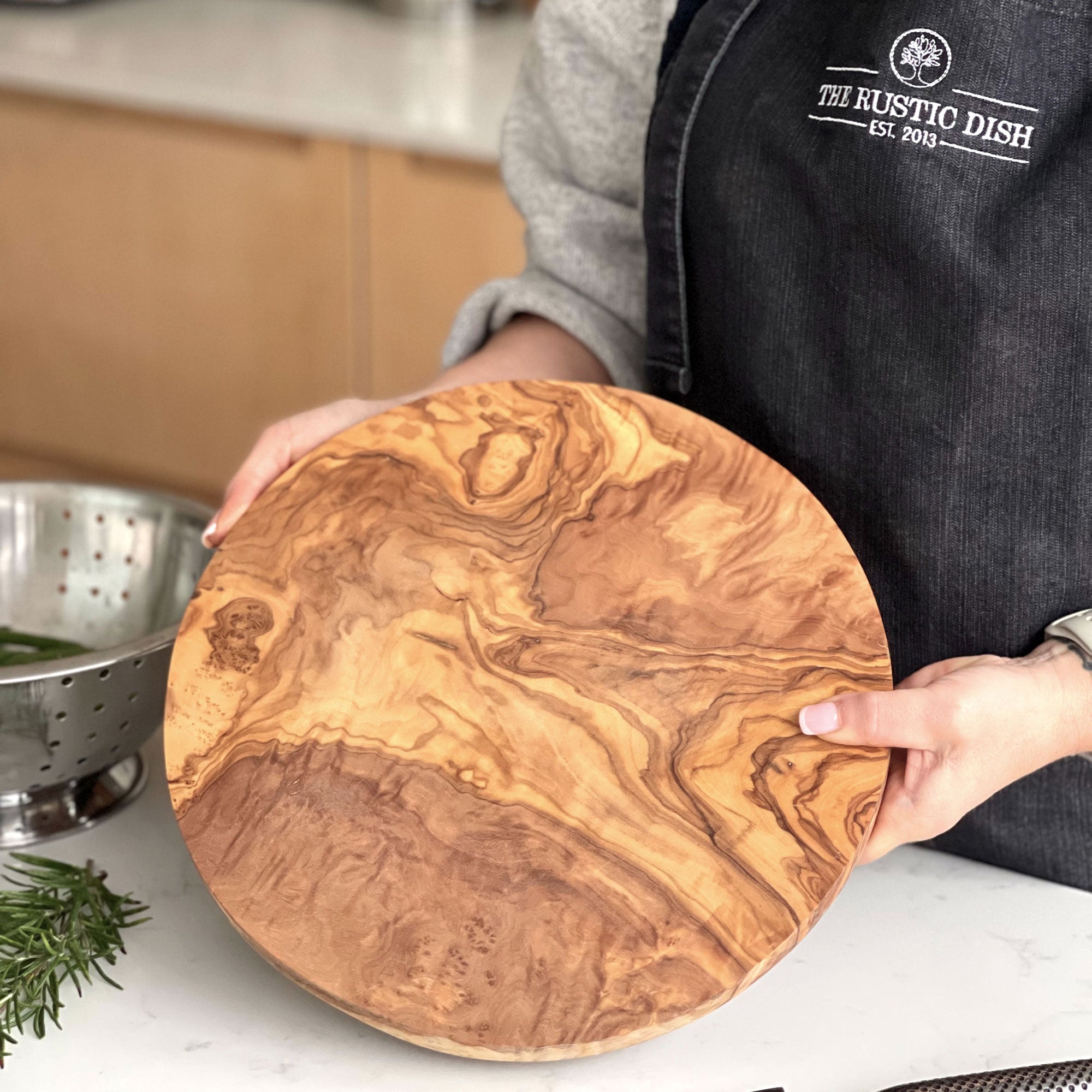 A person holding a round olive wood chopping board with beautiful grain patterns.