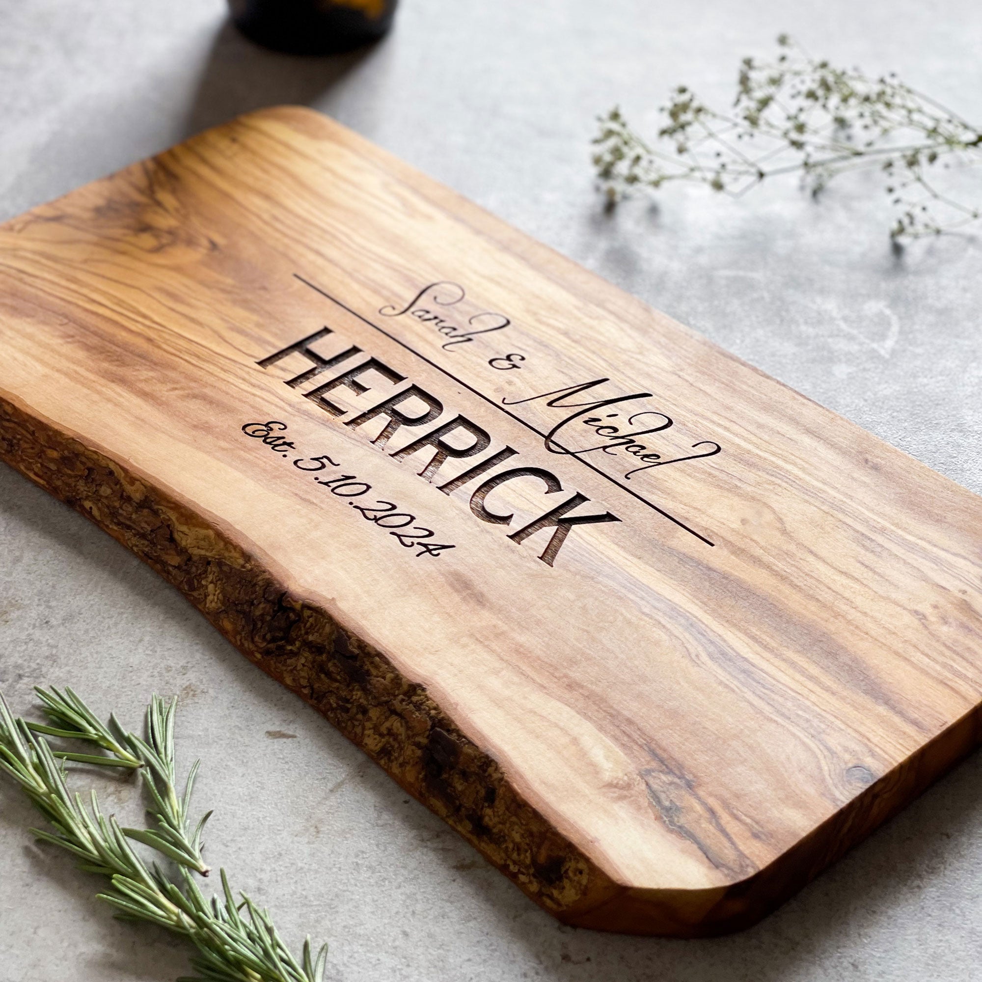 A personalised rustic wooden cheese/chopping board with engraved text, displayed on a grey background with herbs and decorative items.