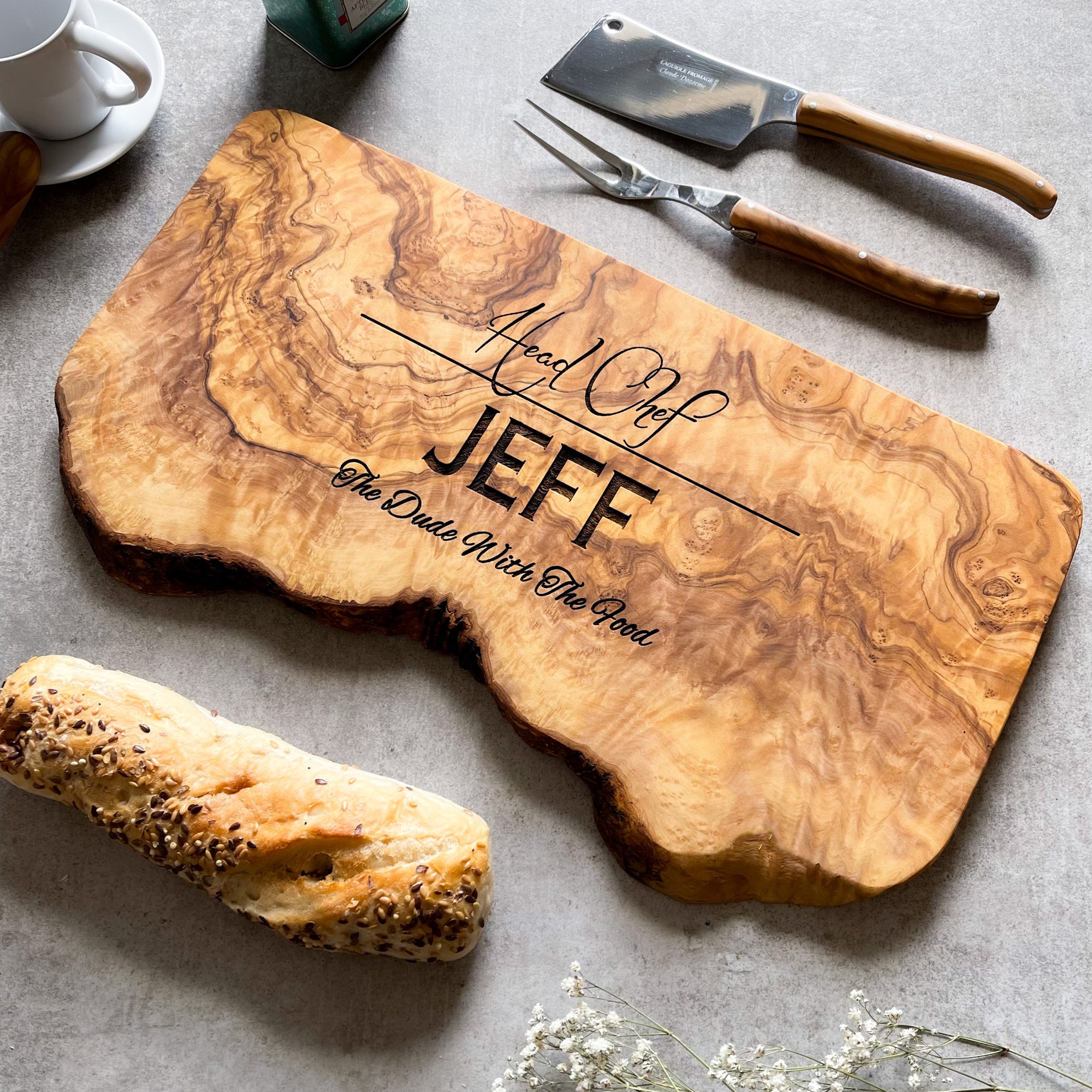 Large personalised olive wood cutting board with engraved text on a grey surface with bread and serving or carving utensils.