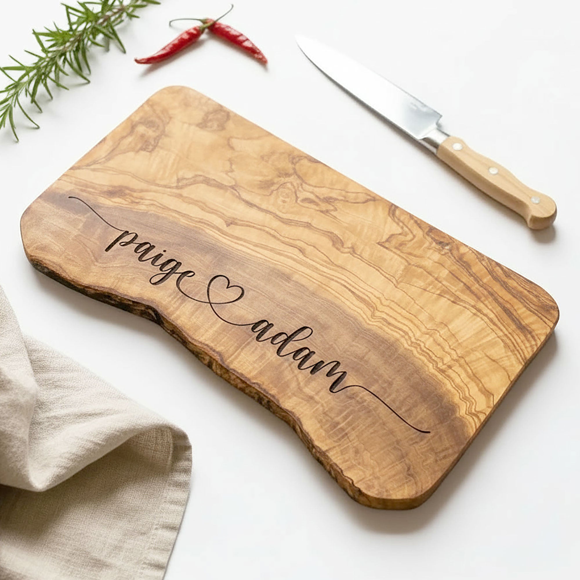 Wooden cutting board with engraved names 'Paige' and 'Adam', knife, and herbs on a white background
