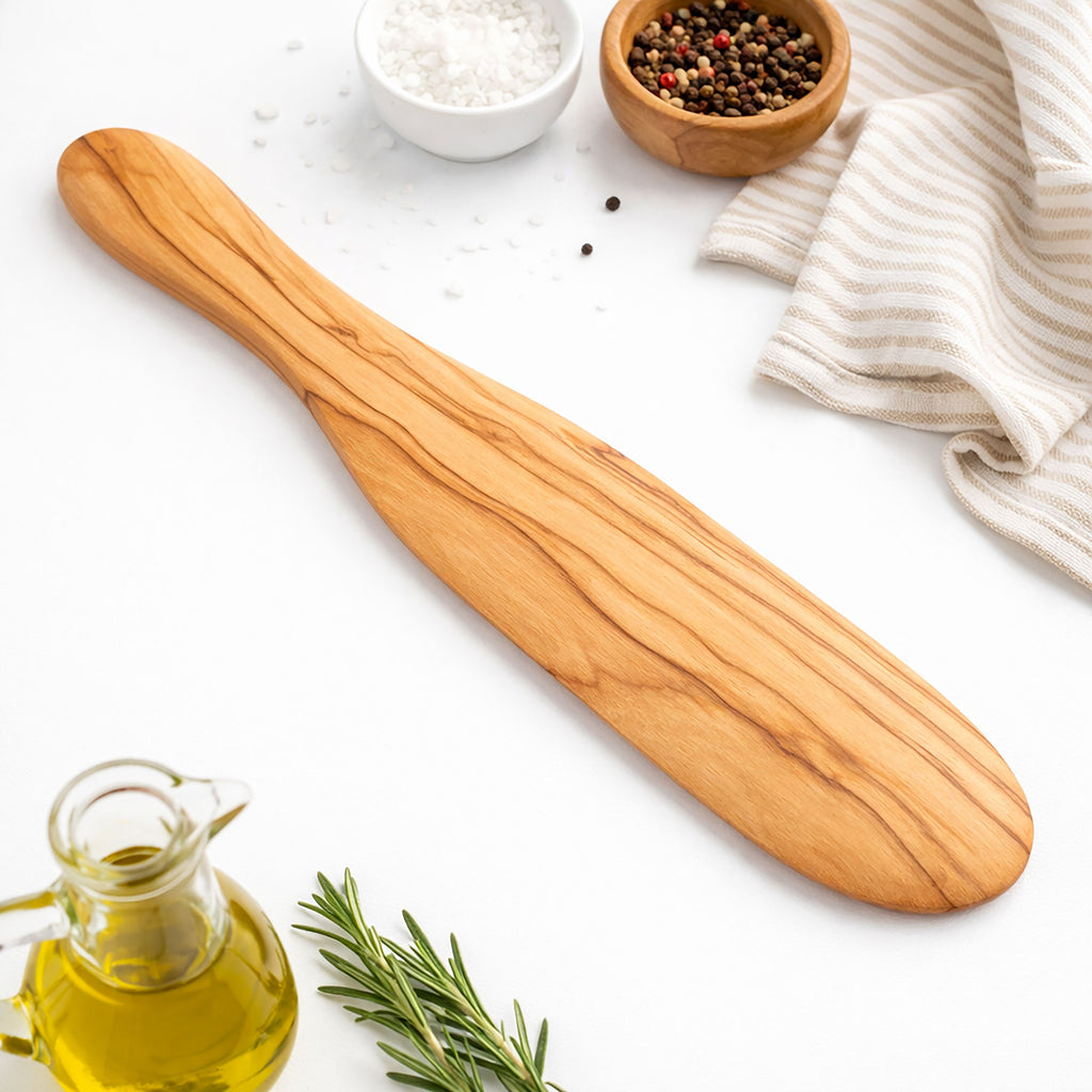 Wooden spatula on a white surface with olive oil, rosemary, and small bowls of salt and pepper.
