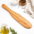 Wooden spatula on a white surface with olive oil, rosemary, and small bowls of salt and pepper.