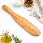 Wooden spatula on a white surface with olive oil, rosemary, and small bowls of salt and pepper.