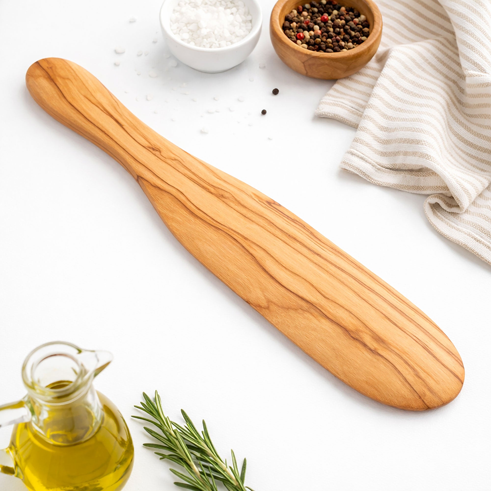 Wooden spatula on a white surface with olive oil, rosemary, and small bowls of salt and pepper.