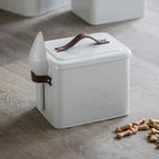 White pet food container with brown handles on a wooden floor with scattered dog treats.
