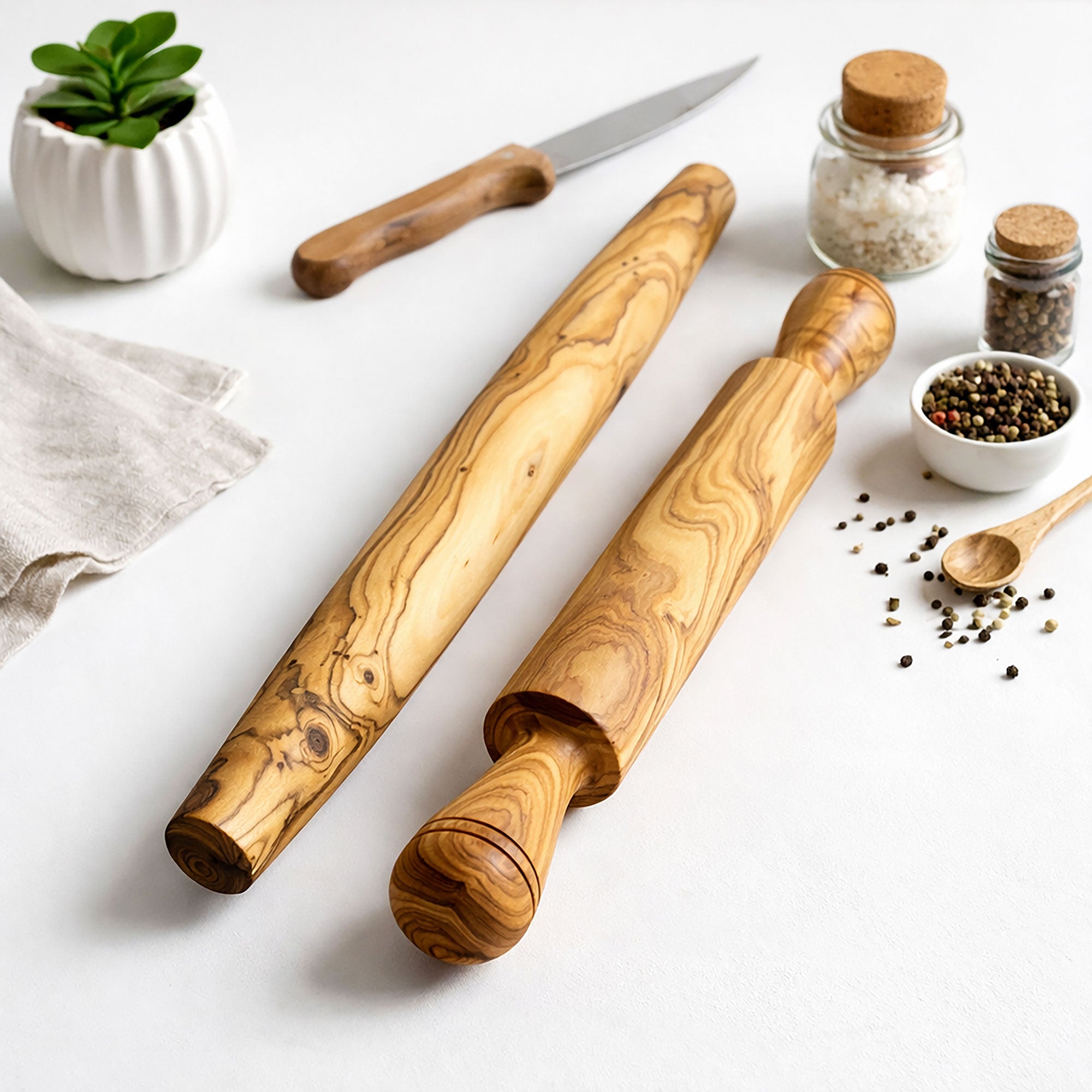 Two wooden rolling pins on a white surface with kitchen items like a knife, small bowls, and a plant.