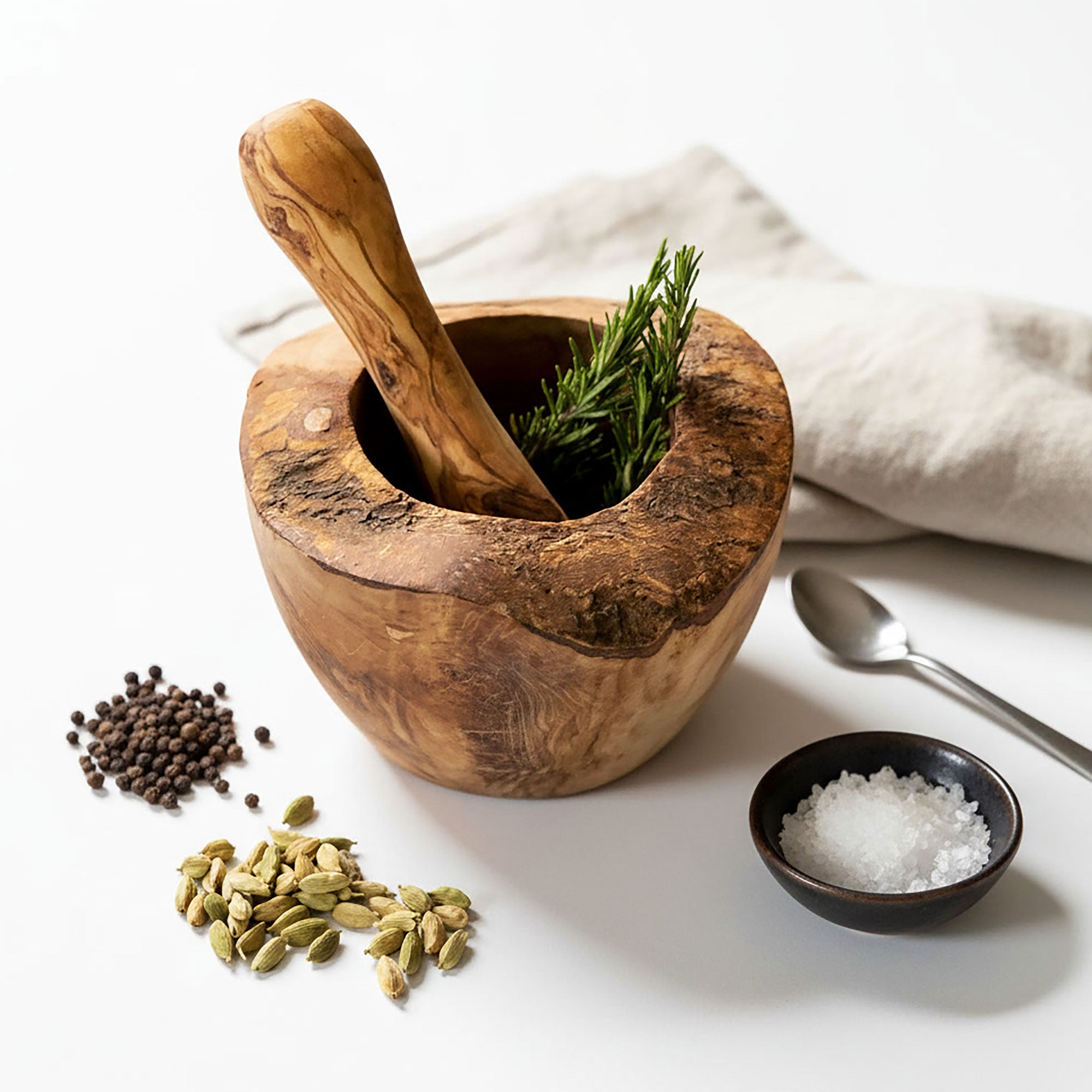 Wooden mortar and pestle with herbs, surrounded by spices and a spoon on a white surface.