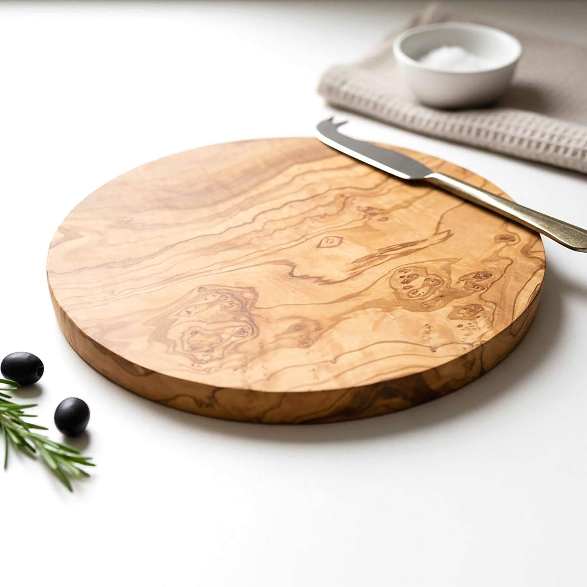 Round wooden cutting board with a knife on a white surface, surrounded by olives and rosemary.