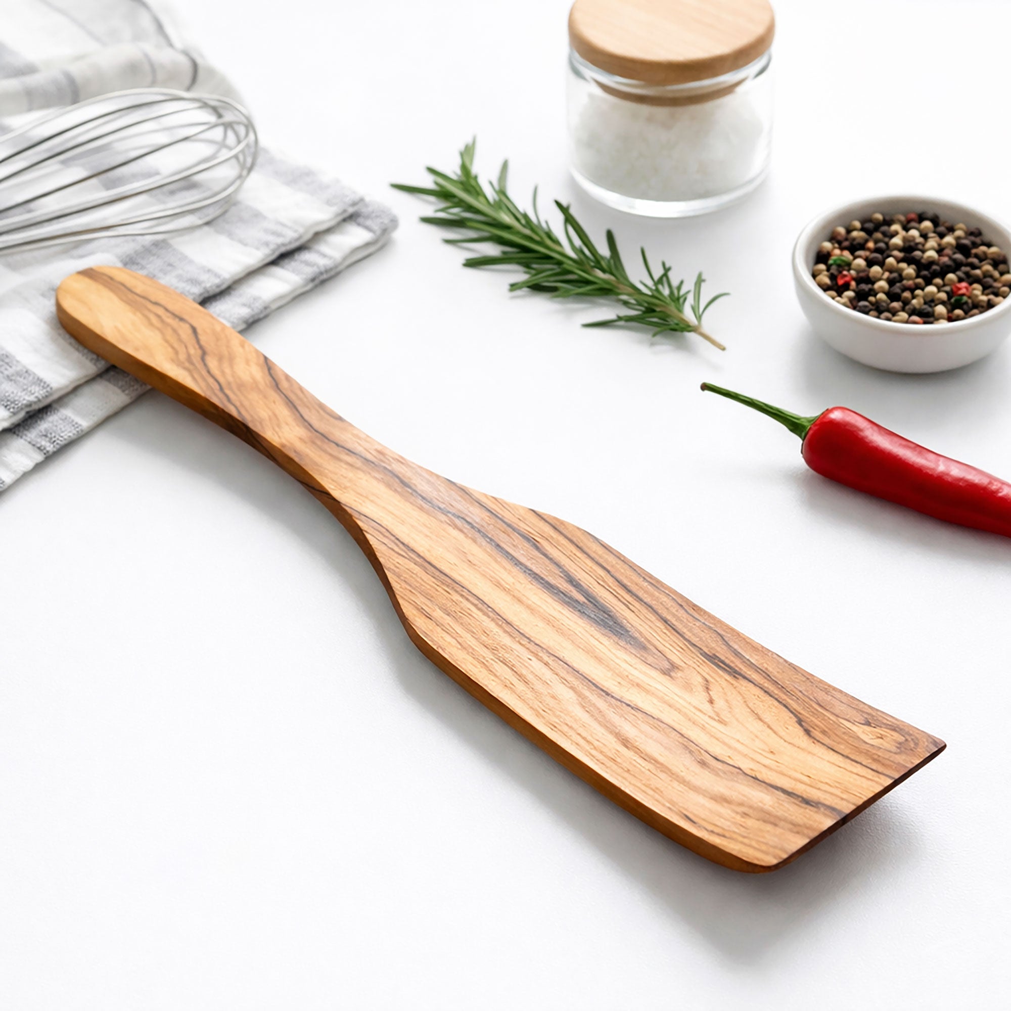 Wooden spatula on a white surface with kitchen items like a whisk, salt shaker, and red pepper.
