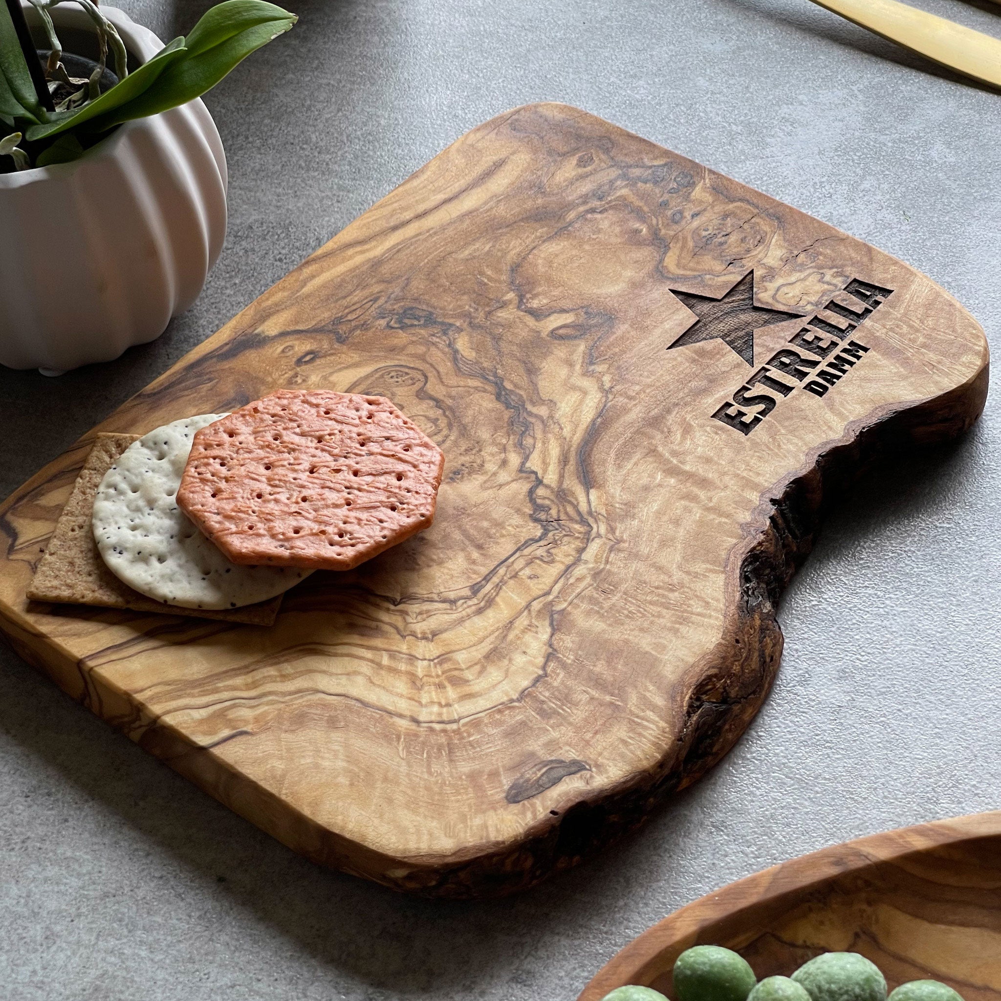 An olive wood chopping/cheese board with a branded logo engraved on it, displayed on a table with some food items.