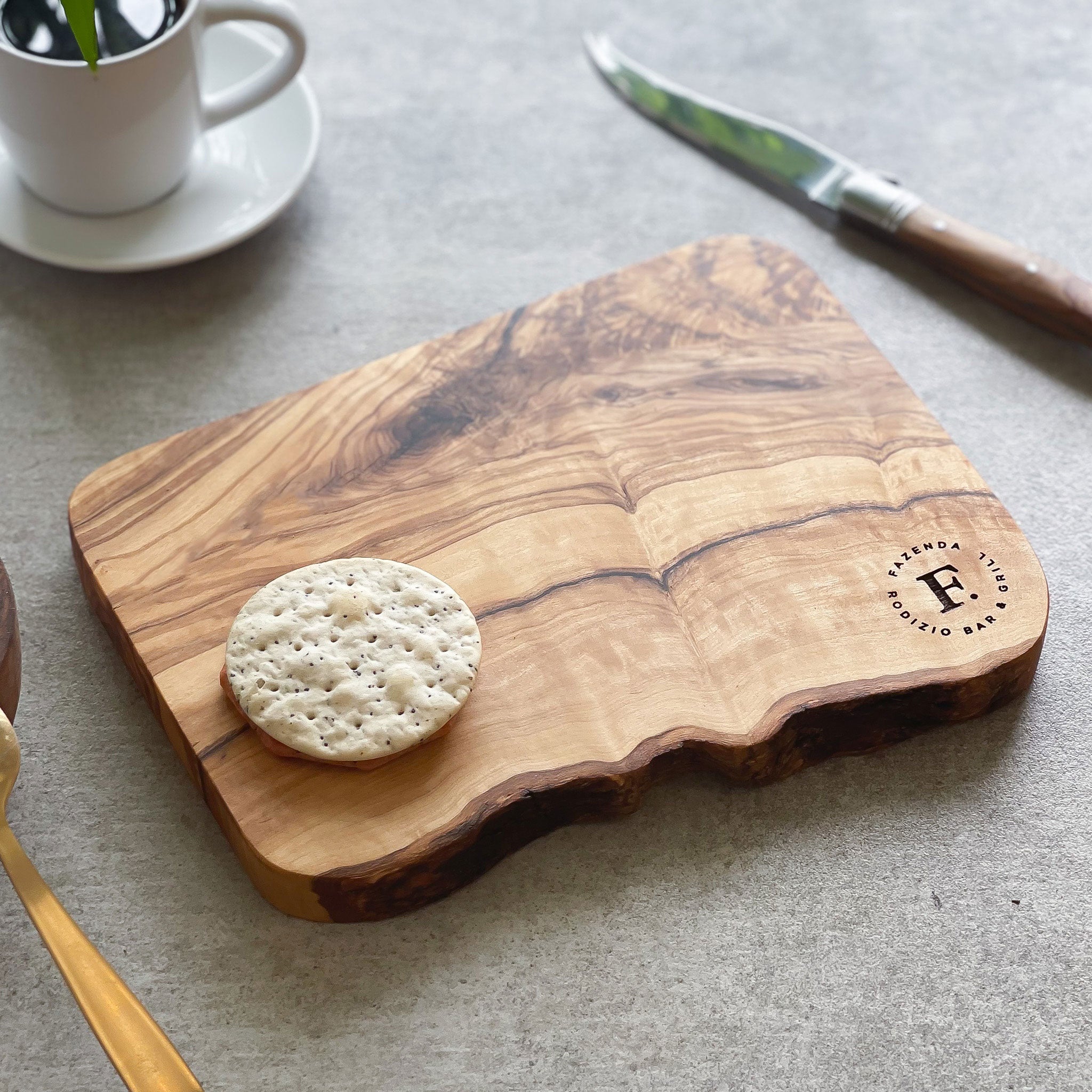 An olive wood chopping/cheese board with a branded logo engraved on it, displayed on a table with some food items.