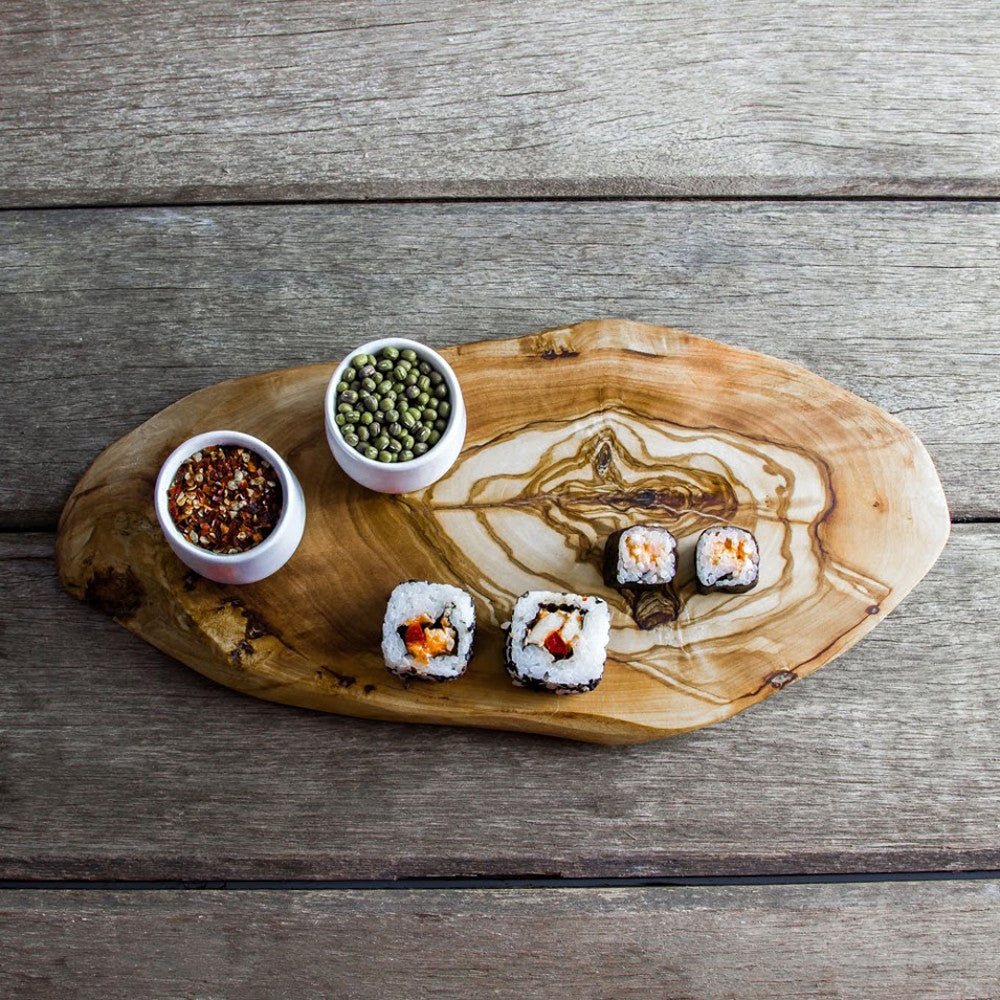 an olive wood cutting board on a grey table surhface with some sushi and seed pots.