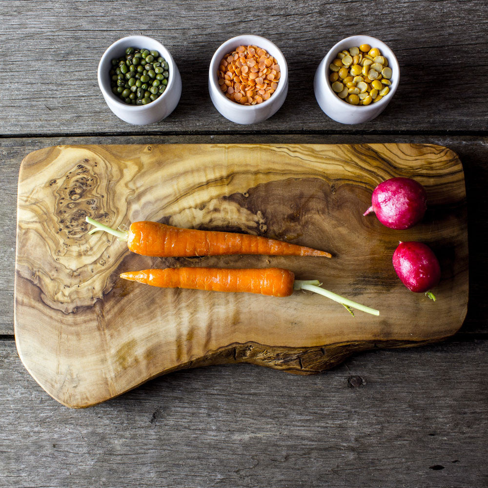 A rustic olive wood cheese/chopping board on a dark wooden surface topped with 2 carrots and 2 beetroots. Alongside the board is 3 small pots containing variety of grains 