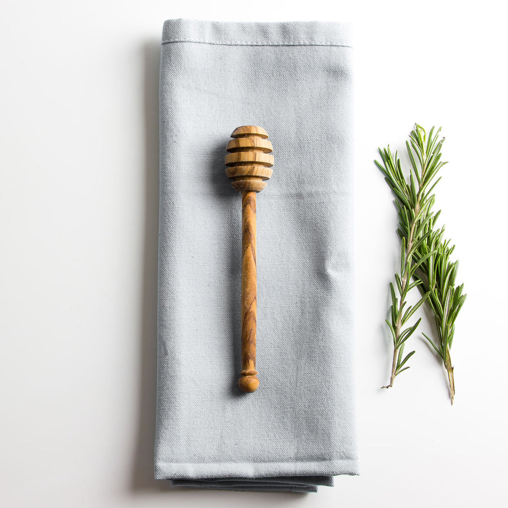 an olive wood honey drizzler resting on a grey napkin with some herbs parallel