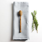 an olive wood honey drizzler resting on a grey napkin with some herbs parallel