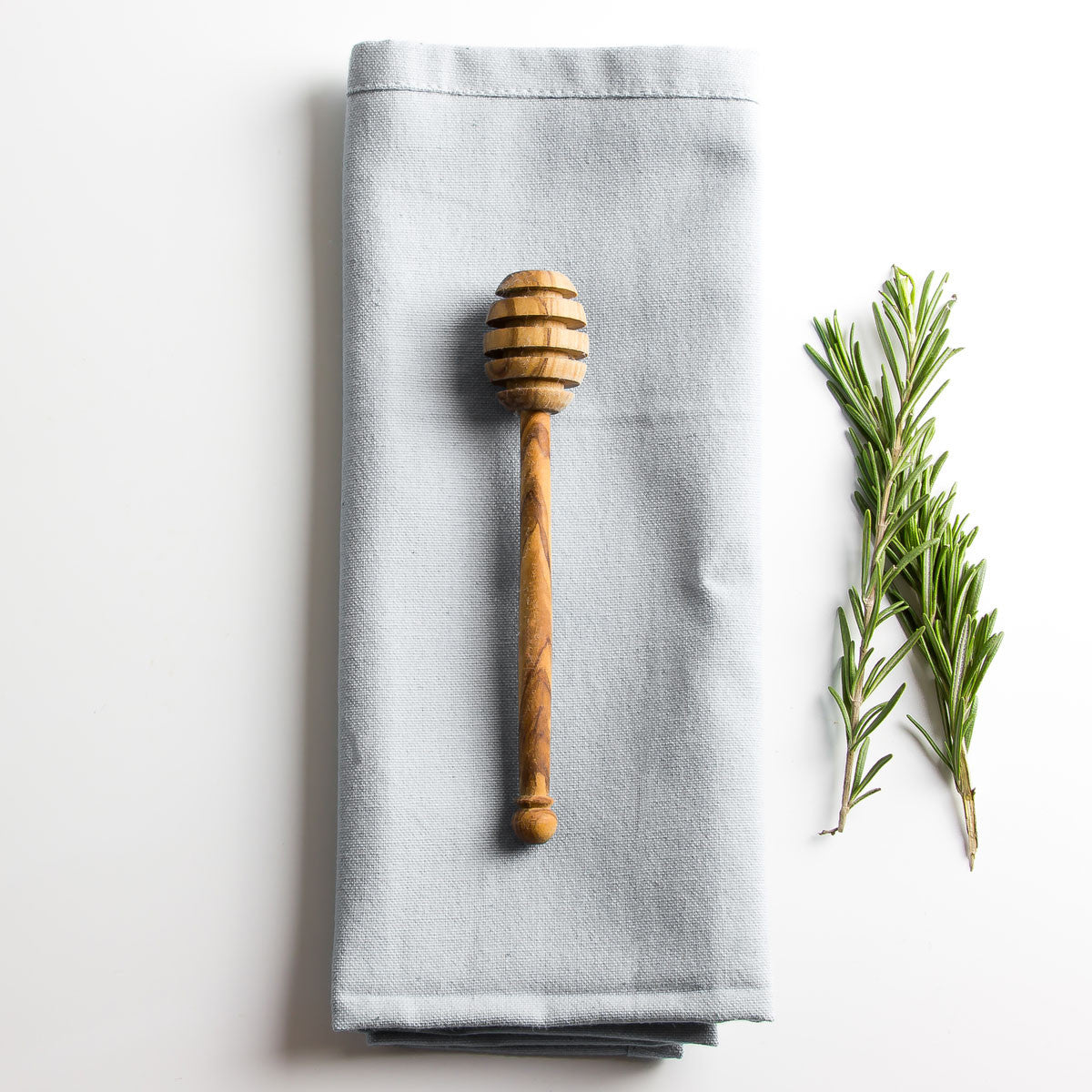 an olive wood honey drizzler resting on a grey napkin with some herbs parallel