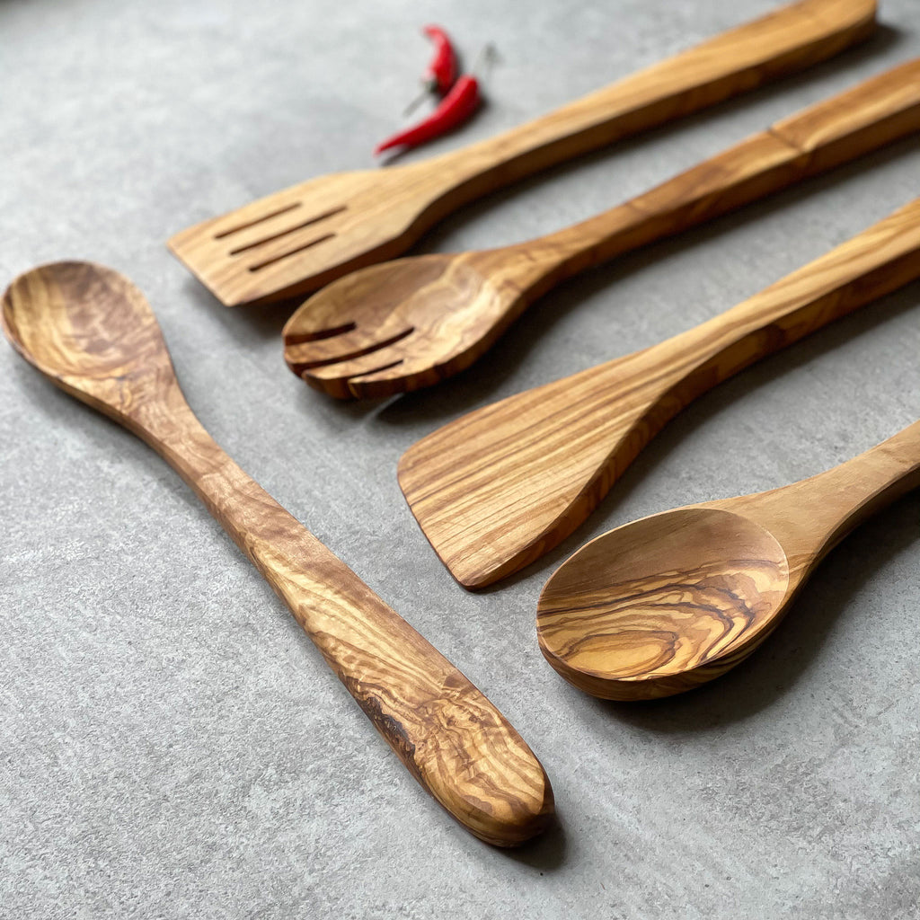 A set of olive wood kitchen utensils including a ladle, slotted spoon, solid spoon, and turner, displayed on a grey surface with a red chili in the background.
