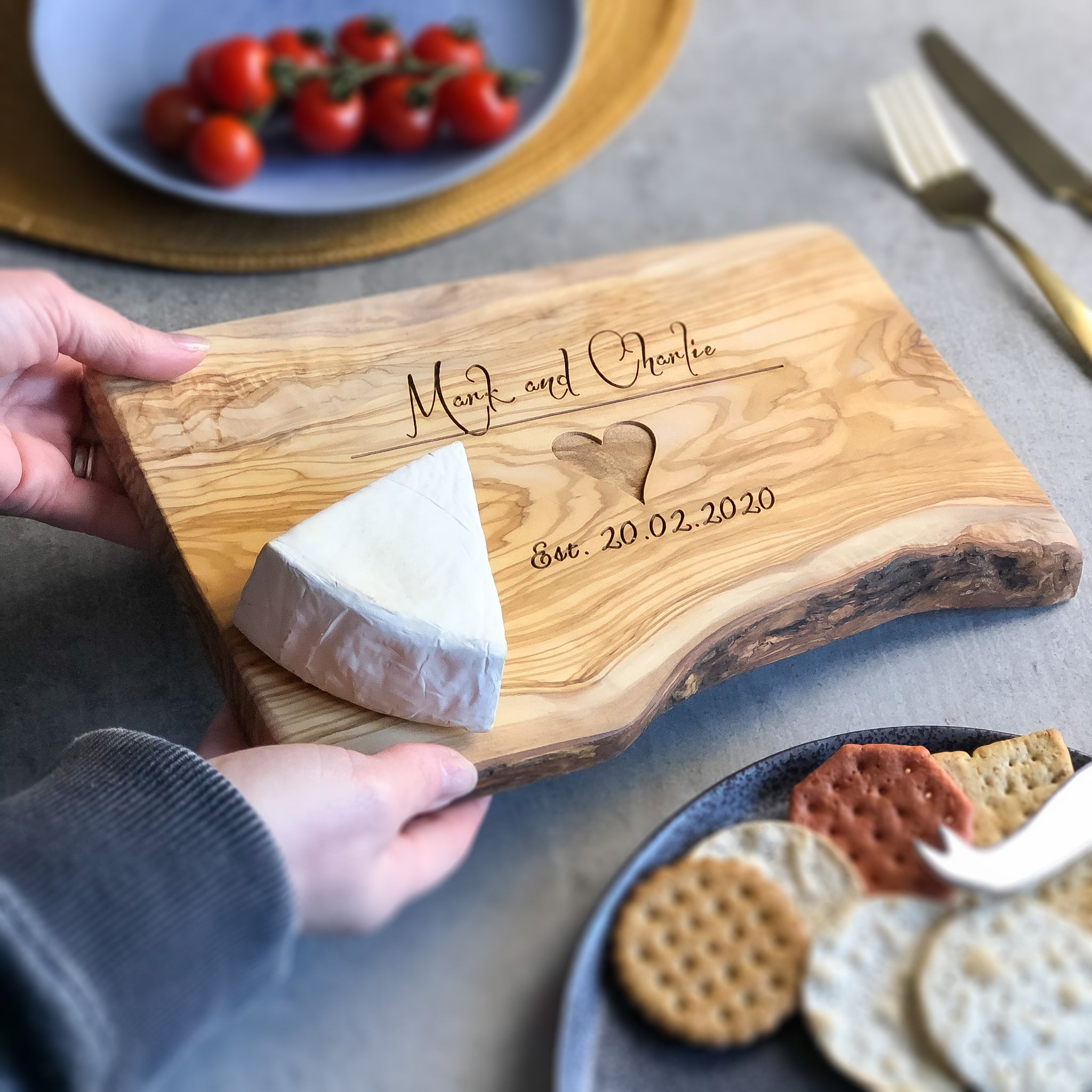 Wooden cutting board with engraved text being placed on a grey surface with cheese and cracker related items.