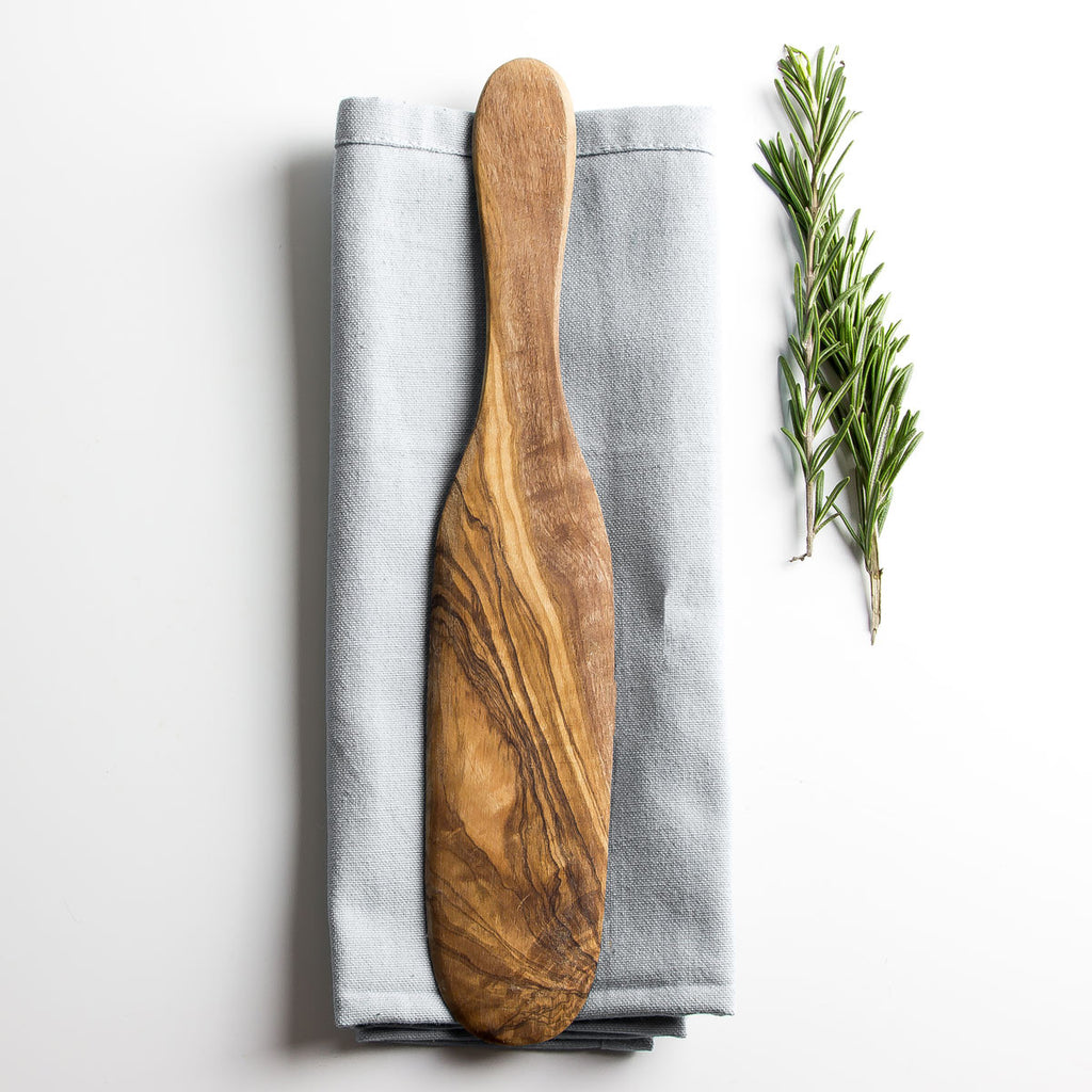olive wood spatula resting on a grey napkin on a plain white surface with some herbs alongside