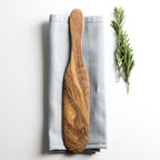 olive wood spatula resting on a grey napkin on a plain white surface with some herbs alongside